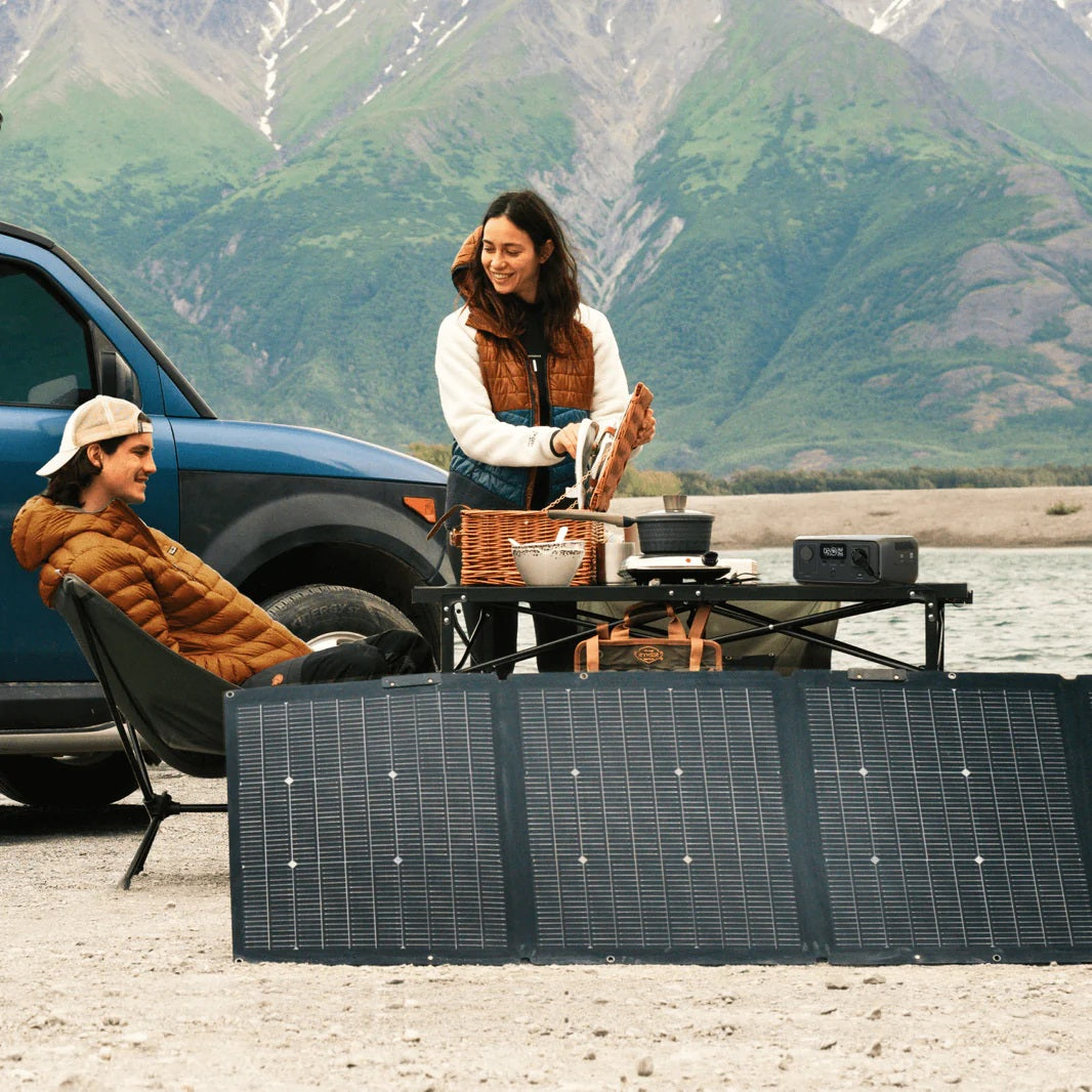 Outdoor camping scene with EcoFlow RIVER 3 portable power station powering devices near solar panels and two people enjoying nature