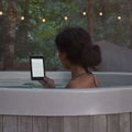 Person relaxing in a hot tub reading a Wireless E-Ink eReader in a serene outdoor setting